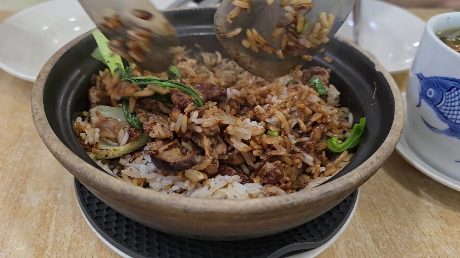 Claypot rice being turned over with two spatulas, the crispy rice from the bottom mixed through, with a blue-and-white soup pot beside it