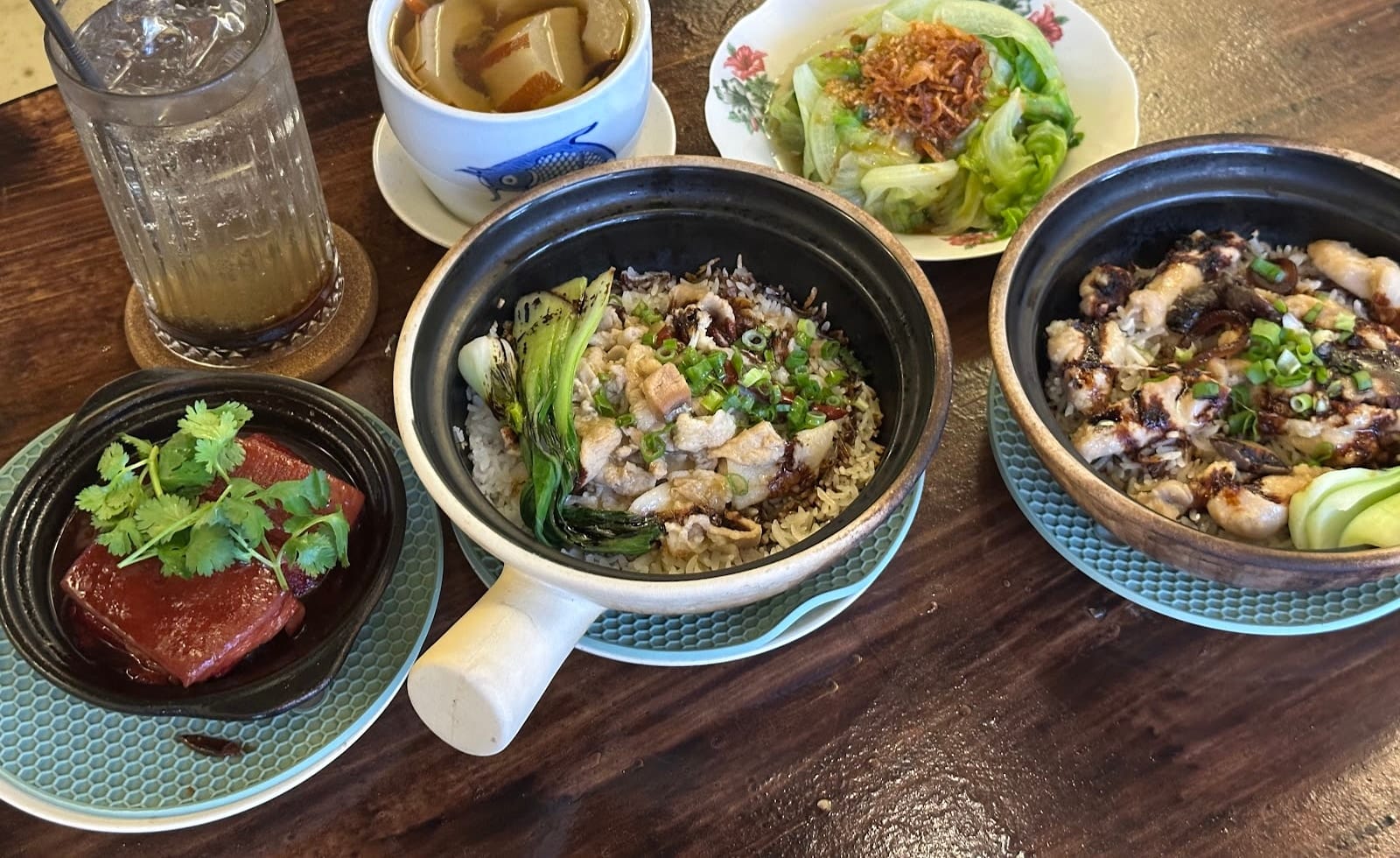 A table with two claypot rice dishes, a pot of pork belly, a bowl of soup, greens and a drink