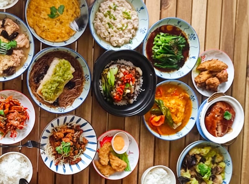 An overhead view of a table of claypot rice, steamed fish, omelette, vegetables, fried chicken and soups in blue-and-white bowls